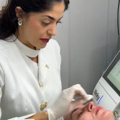 Dra. Thamara Martins, de jaleco branco e luvas, fazendo limpeza de pele em uma paciente deitada na maca